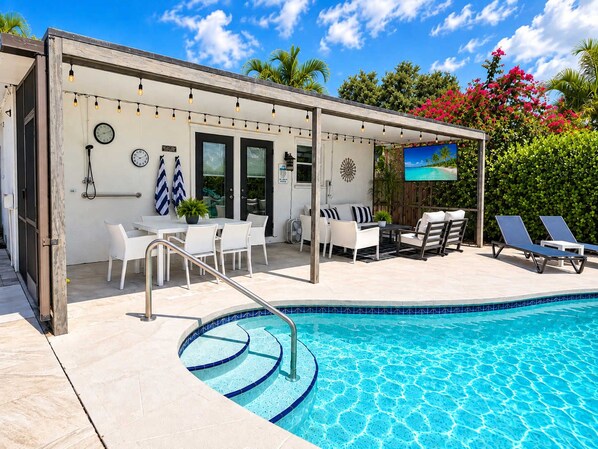 A covered outdoor area with a pool, seating, and a television screen.