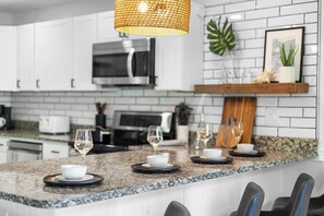 Una cocina moderna con gabinetes blancos, encimera de granito, lámpara colgante y estante de madera con objetos decorativos.
