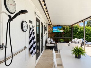 A covered outdoor seating area with a television screen displaying a tropical beach scene, a clock, and a towel rack.