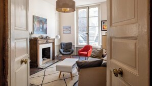 A room with a fireplace, a sofa, a red armchair, and a large window with a view of buildings.