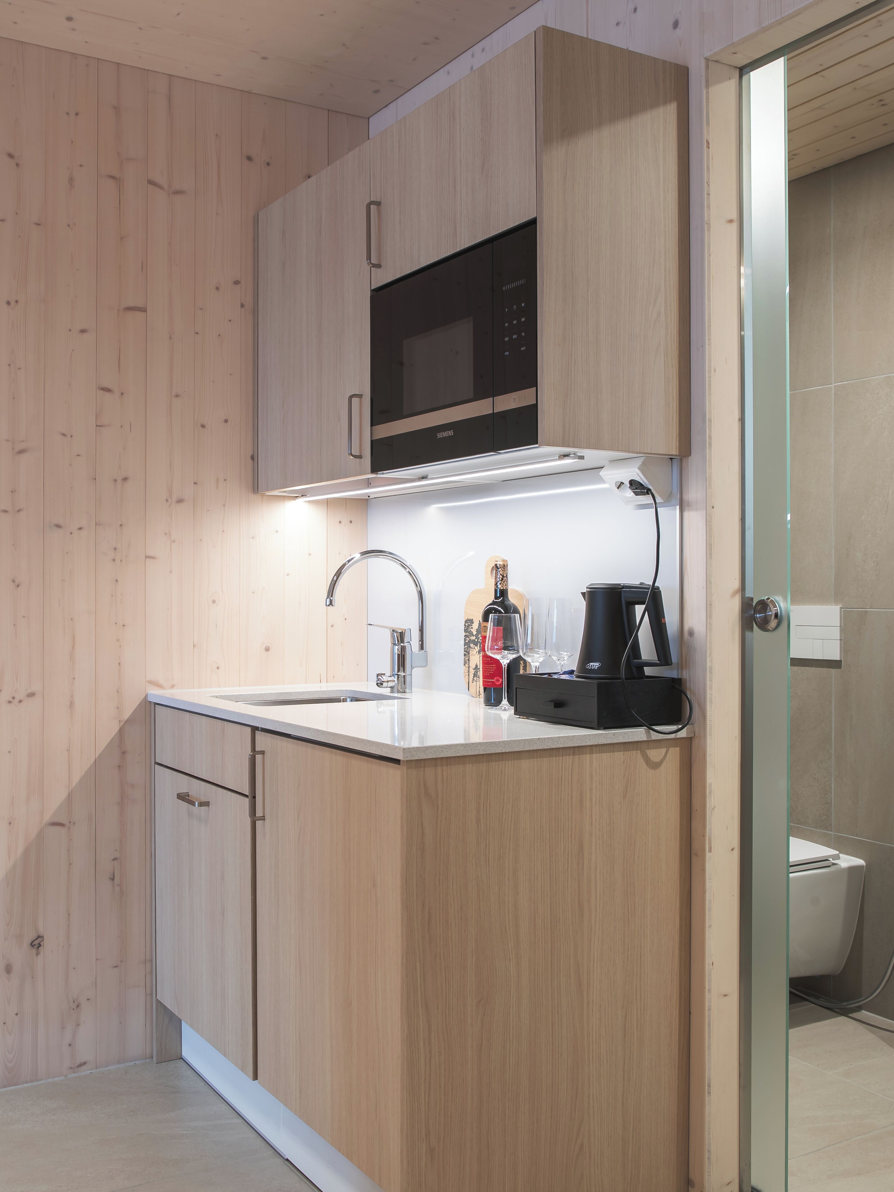 Una cocina moderna con armarios de madera, microondas integrado, cafetera y cabina de ducha de vidrio.