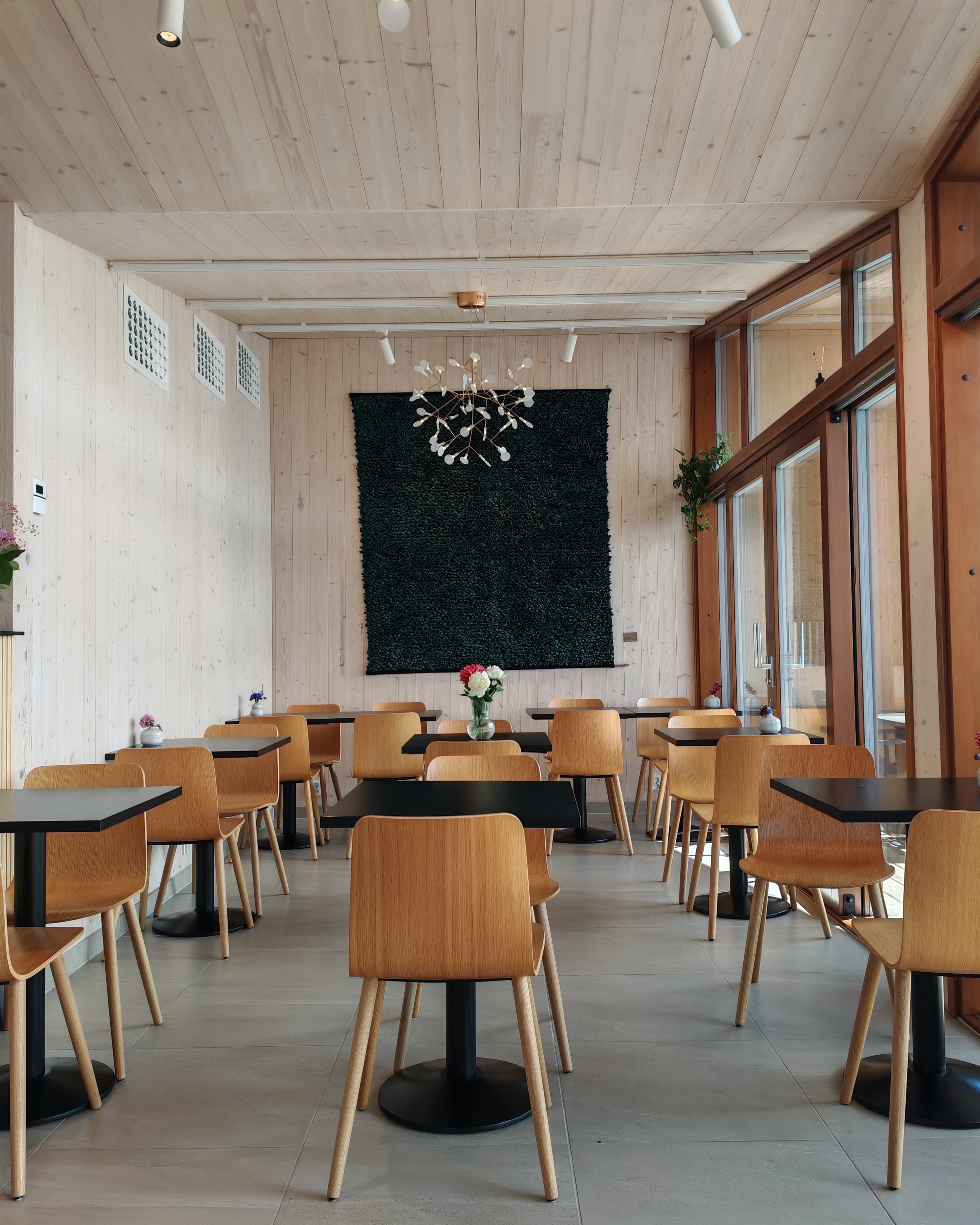A dining area with wooden tables and chairs, a green wall with a decorative light fixture, and large windows.