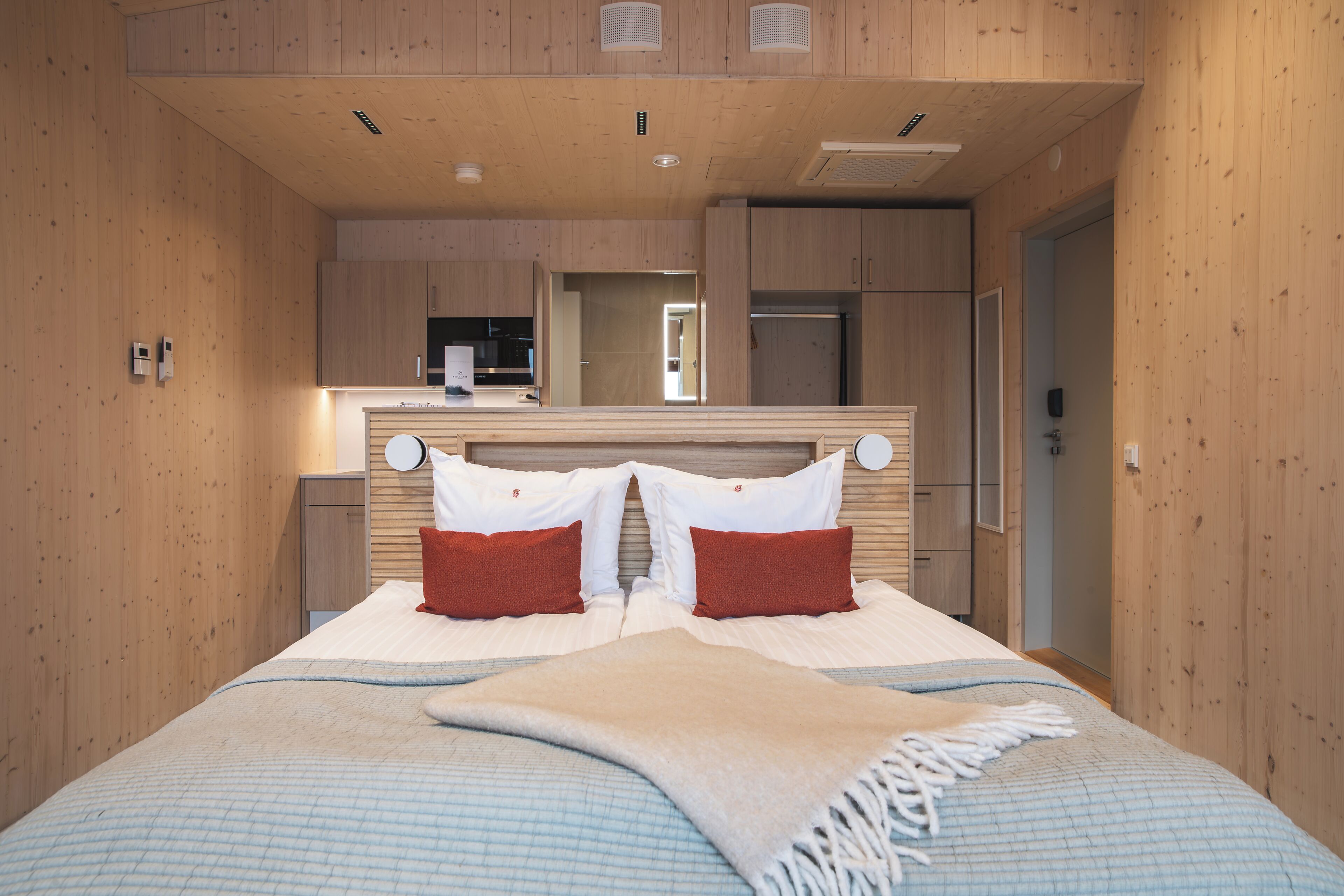 A bedroom with a bed, pillows, a blanket, a wooden ceiling, and a kitchen area visible through an open door.