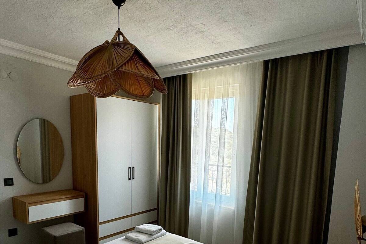 A hotel room with a large bed, a wooden wardrobe, a round mirror, a vanity with a mirror, and a window with curtains.
