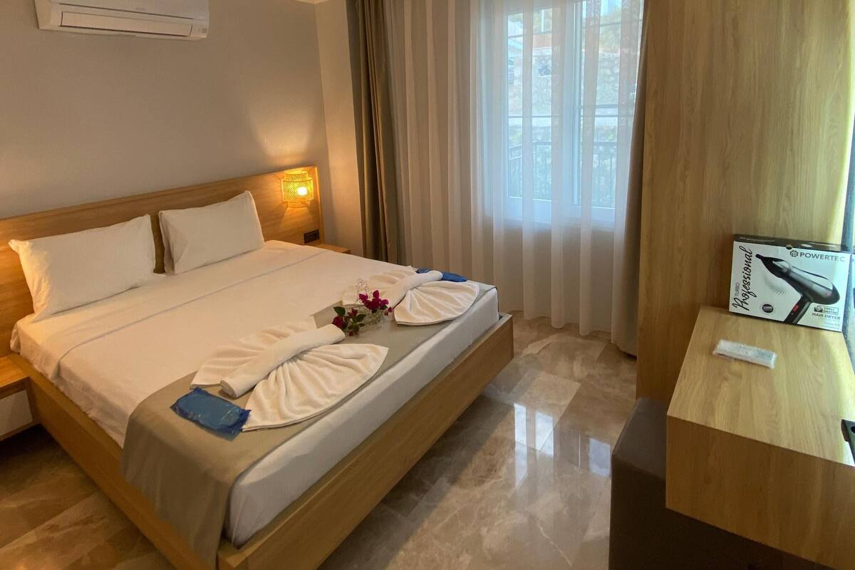 A hotel room with a wooden bed, white linens, a bedside table with a lamp, an air conditioner, and a view of the outdoors through curtains.