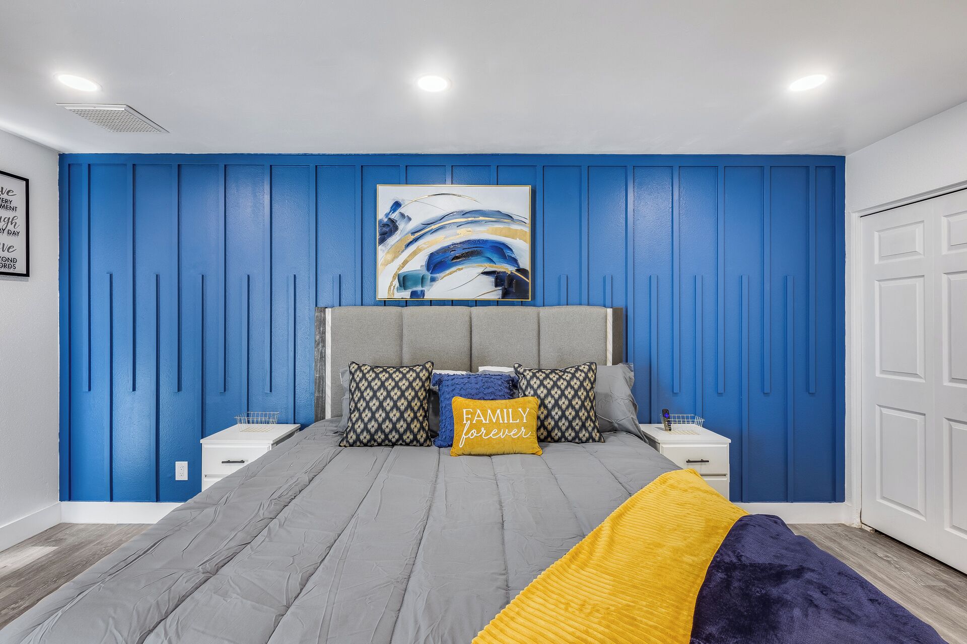 A bedroom with a blue paneled wall, a grey bed with patterned pillows, and a painting above the bed.