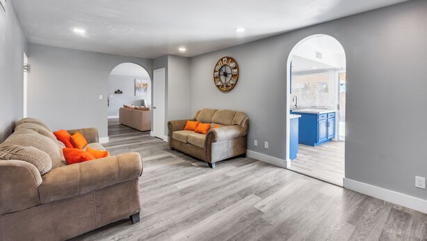A spacious living area with a grey sofa, a smaller sofa with orange cushions, and a wooden coffee table. A large wall clock and an arched doorway lead to a kitchen area. - Charming 3-bedroom house with AC, WiFi in fabulous Glendale (Glendale)