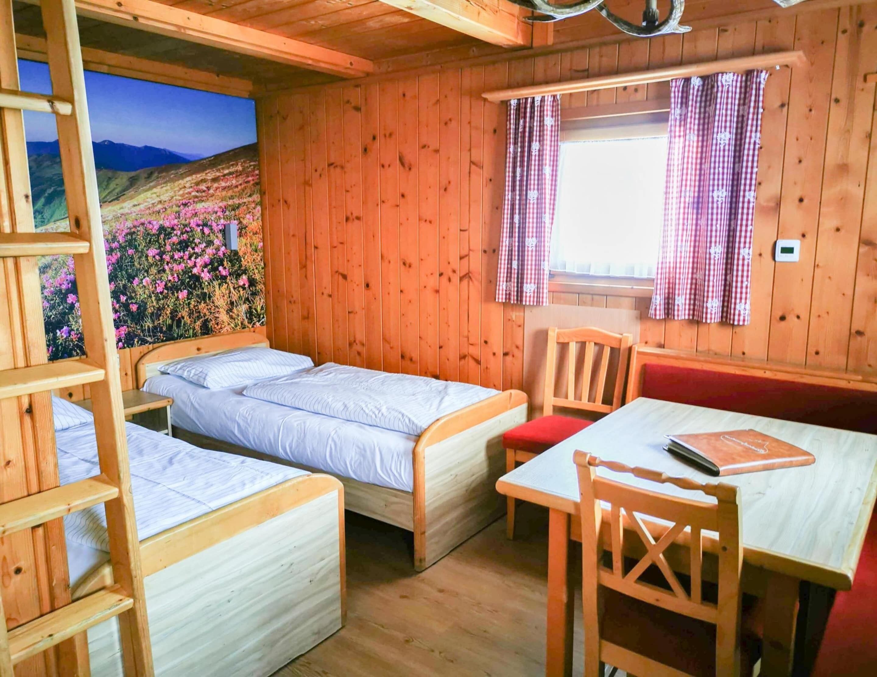 A cabin with two single beds, a wooden table, a bench, and a view of a flower-covered hillside.