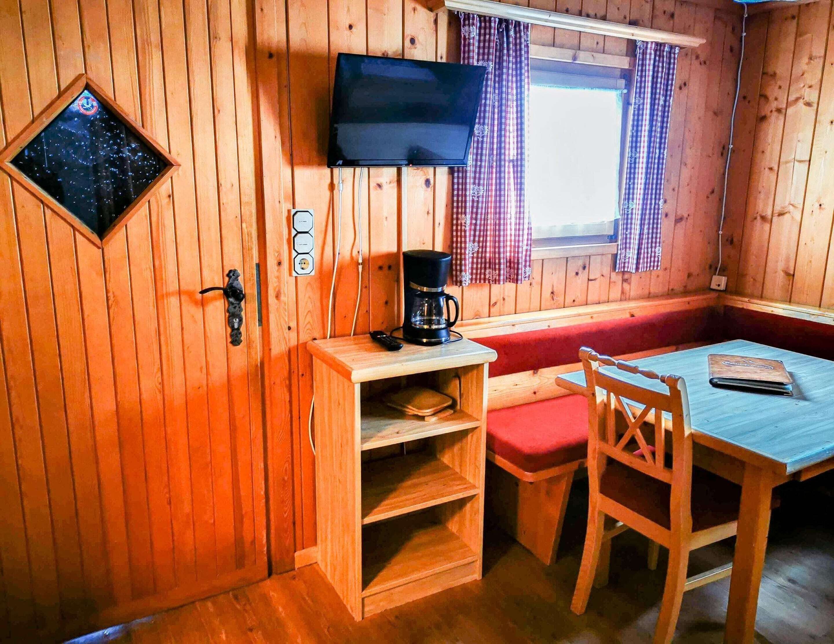 A cozy cabin interior with a wooden wall, a television mounted above a window with checkered curtains, a coffee maker on a wooden table, and a red cushioned bench.