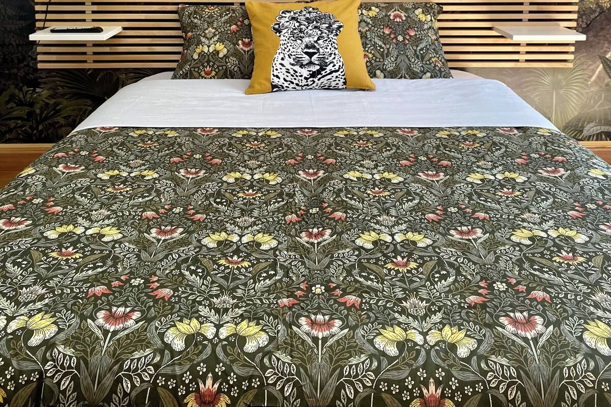 A bedroom with a bed, a wooden headboard, a nightstand with lamps, and a mural of a tropical landscape.