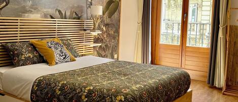 A bedroom with a bed, a mural, wooden flooring, and a view through a window.