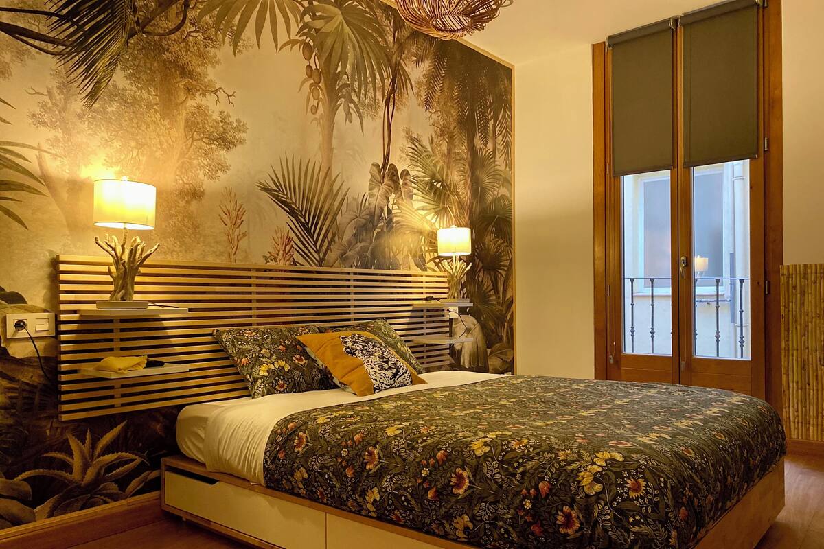 A bedroom with a bed, two bedside lamps, a nightstand, a wall mural, and a sliding door to a balcony.