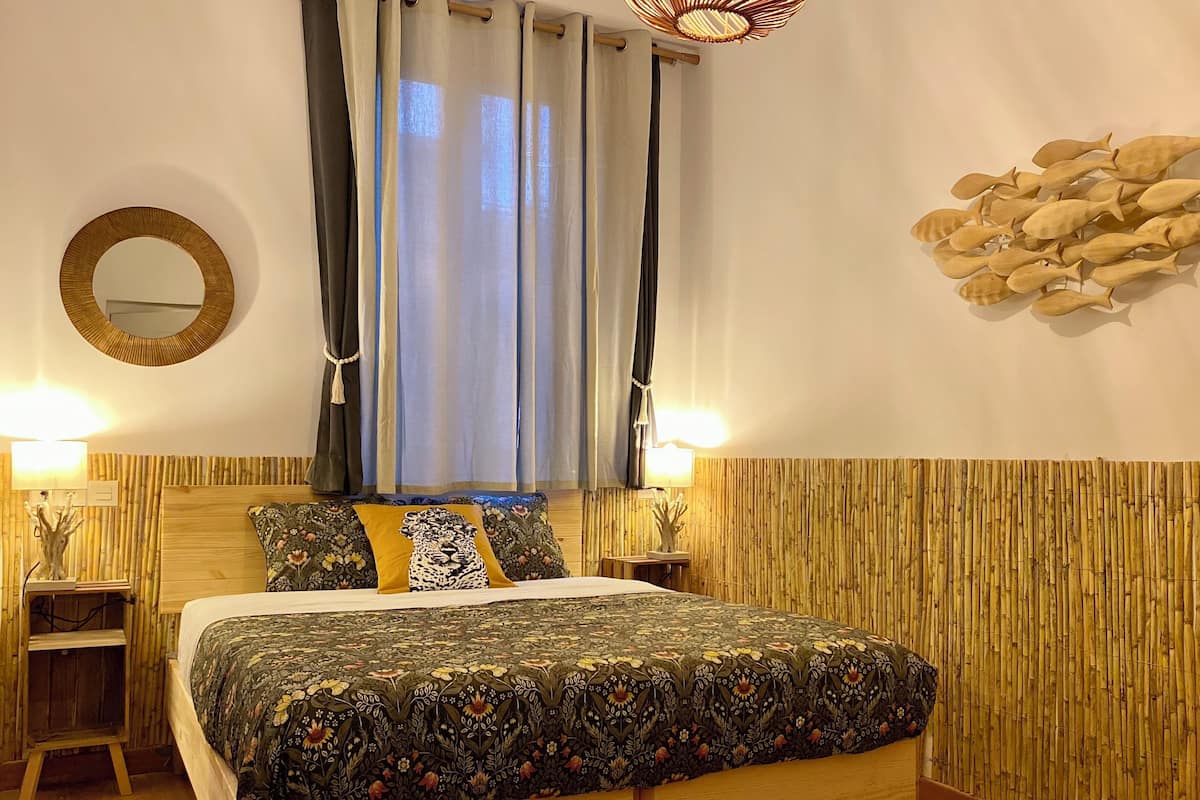 A bedroom with a bed, two bedside tables, a mirror, a window with curtains, and a wall decoration.