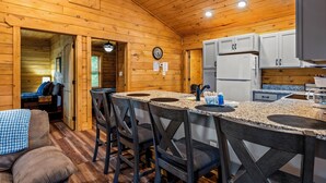 L’intérieur d’une cabane en bois comprend un coin cuisine doté de plans de travail en granit, d’armoires blanches et d’un réfrigérateur. On y trouve également une table à manger avec des chaises et un canapé dans l’espace salon.