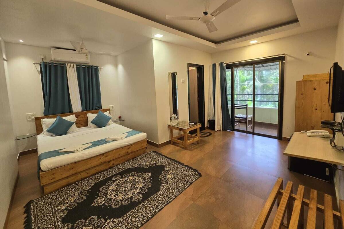 A hotel room with a bed, a desk, a chair, a ceiling fan, and a view through a glass door.