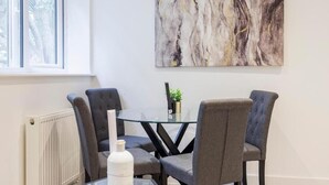 Un comedor con una mesa de vidrio, cuatro sillas tapizadas de color gris, un radiador blanco y un cuadro abstracto grande en la pared.