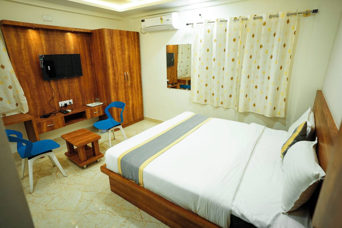 A hotel room with a bed, a desk, a chair, a TV mounted on the wall, and a window with curtains.