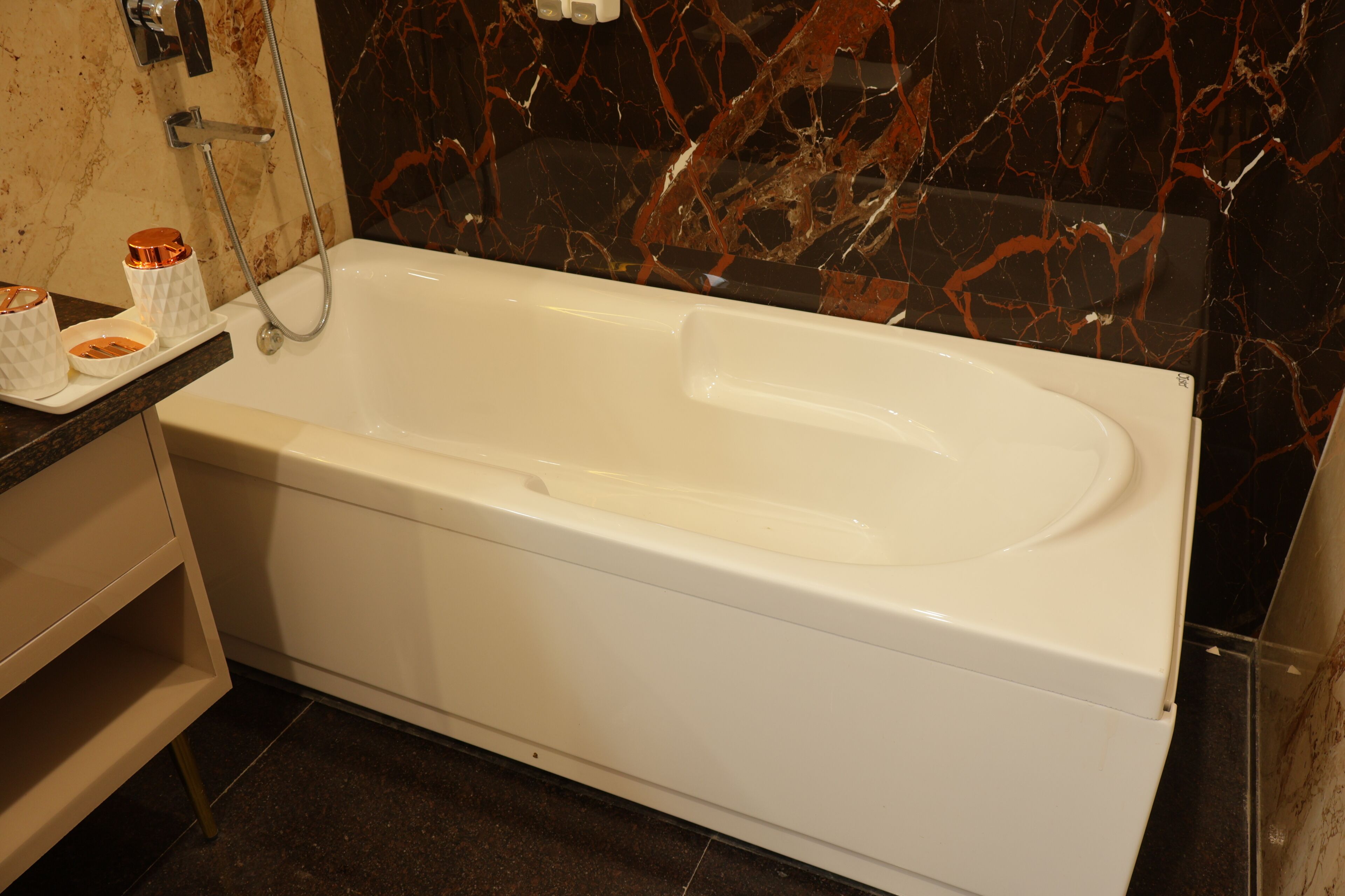 Un baño moderno con una bañera blanca, paredes de mármol y un pequeño estante con artículos de aseo.