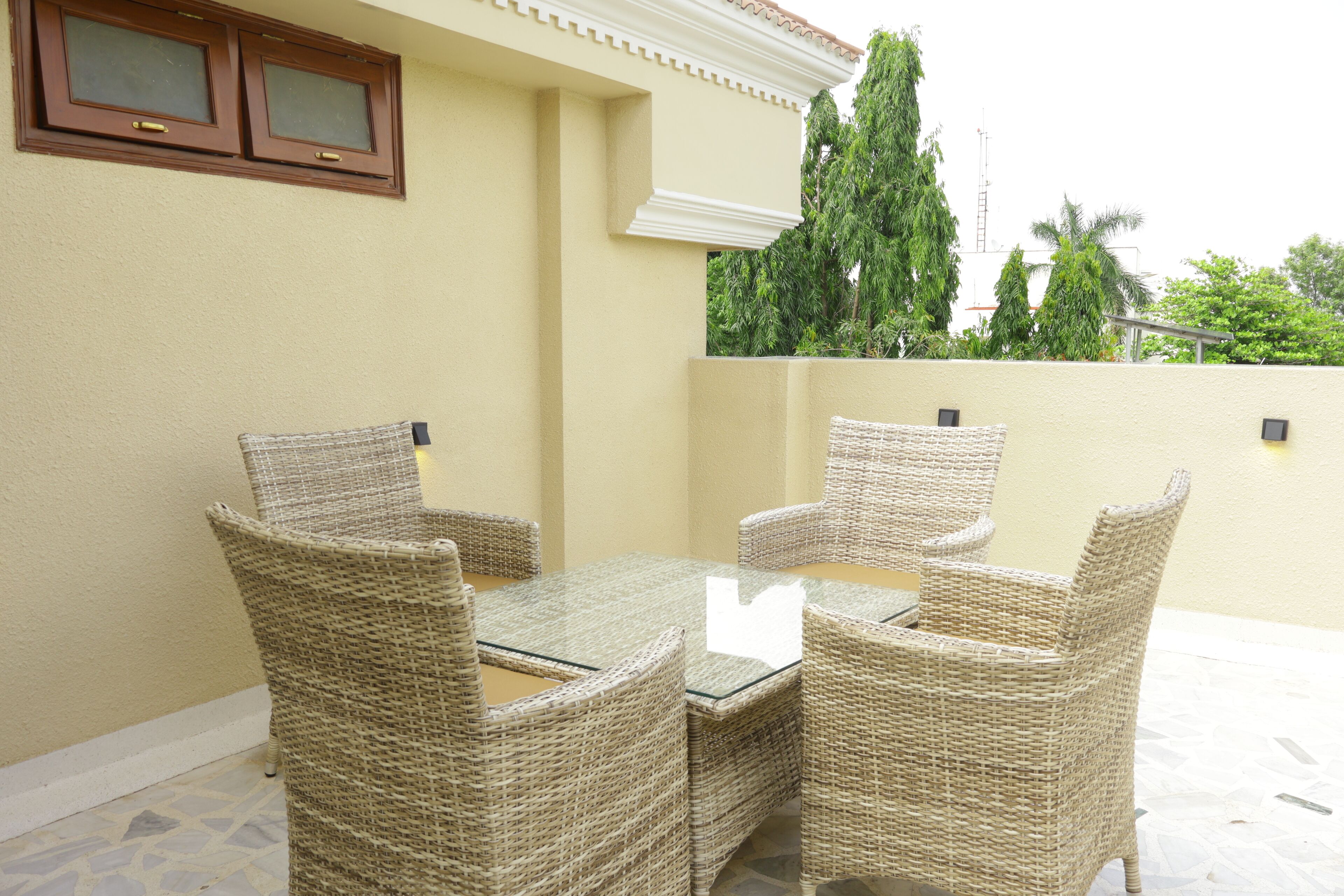 A patio with wicker furniture and a glass table, beige walls, and a view of trees and a building in the background.
