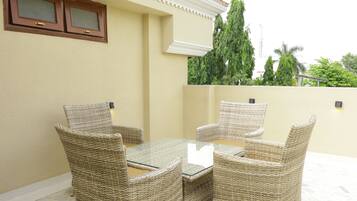 A patio with wicker furniture and a glass table, beige walls, and a view of trees and a building in the background.