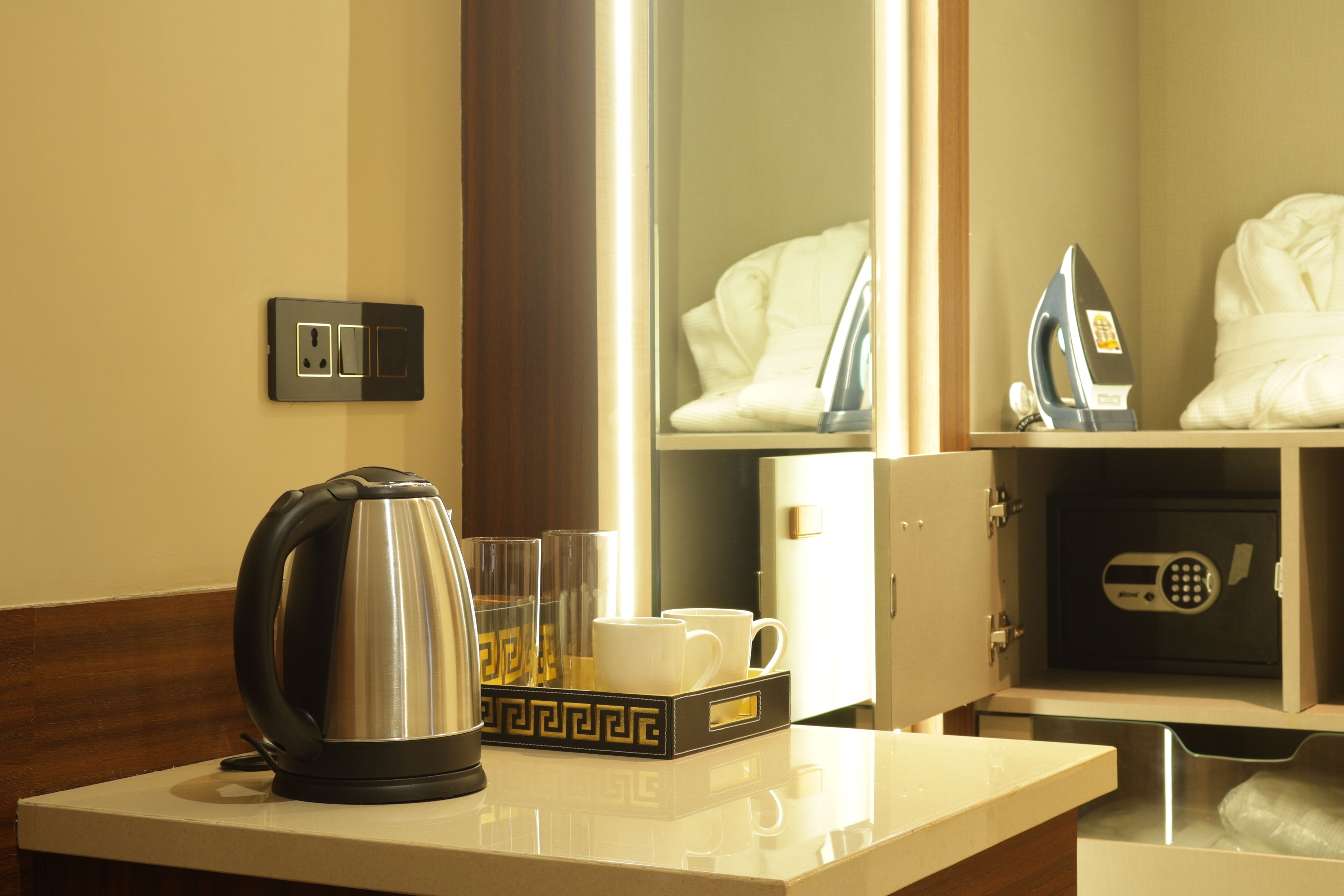 A hotel room with a coffee maker, two cups, a tray, a mirror, an ironing board, and a safe.