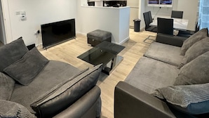 A modern living room with a flat-screen TV, grey sofas, a coffee table, and a kitchen area in the background. - London City Chic Apartment With 3 Bedrooms (London)