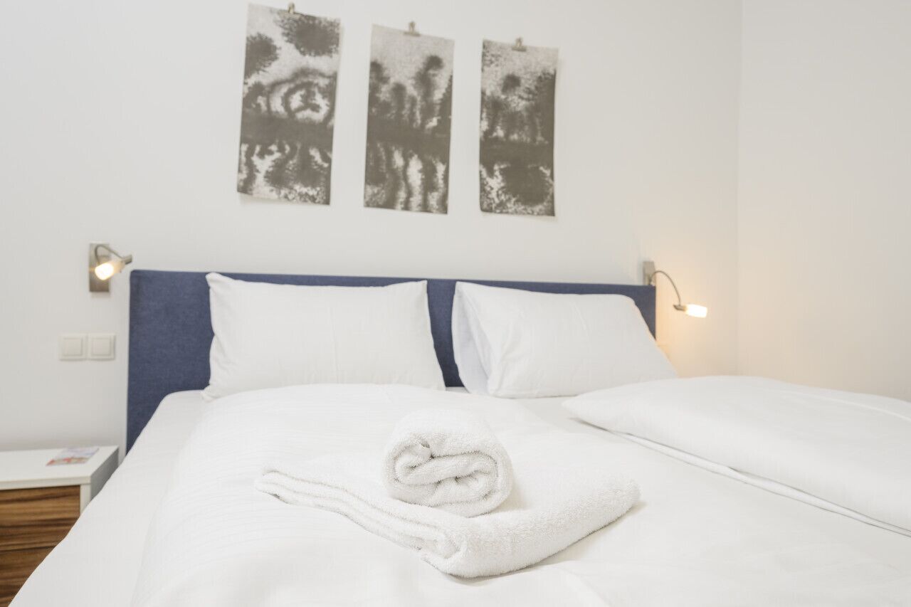 A neatly made bed with white linens and a rolled towel, a blue headboard, and three wall-mounted abstract art pieces.