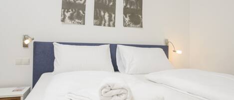 A neatly made bed with white linens and a rolled towel, a blue headboard, and three wall-mounted abstract art pieces.