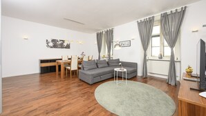 Un salón moderno con un sofá gris, una mesa de comedor de madera y sillas. Hay una alfombra redonda pequeña, un televisor y una ventana con cortinas grises.