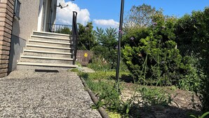 Un edificio con fachada de ladrillo, una escalera exterior que lleva a un balcón y un jardín con un farol y diversas plantas.