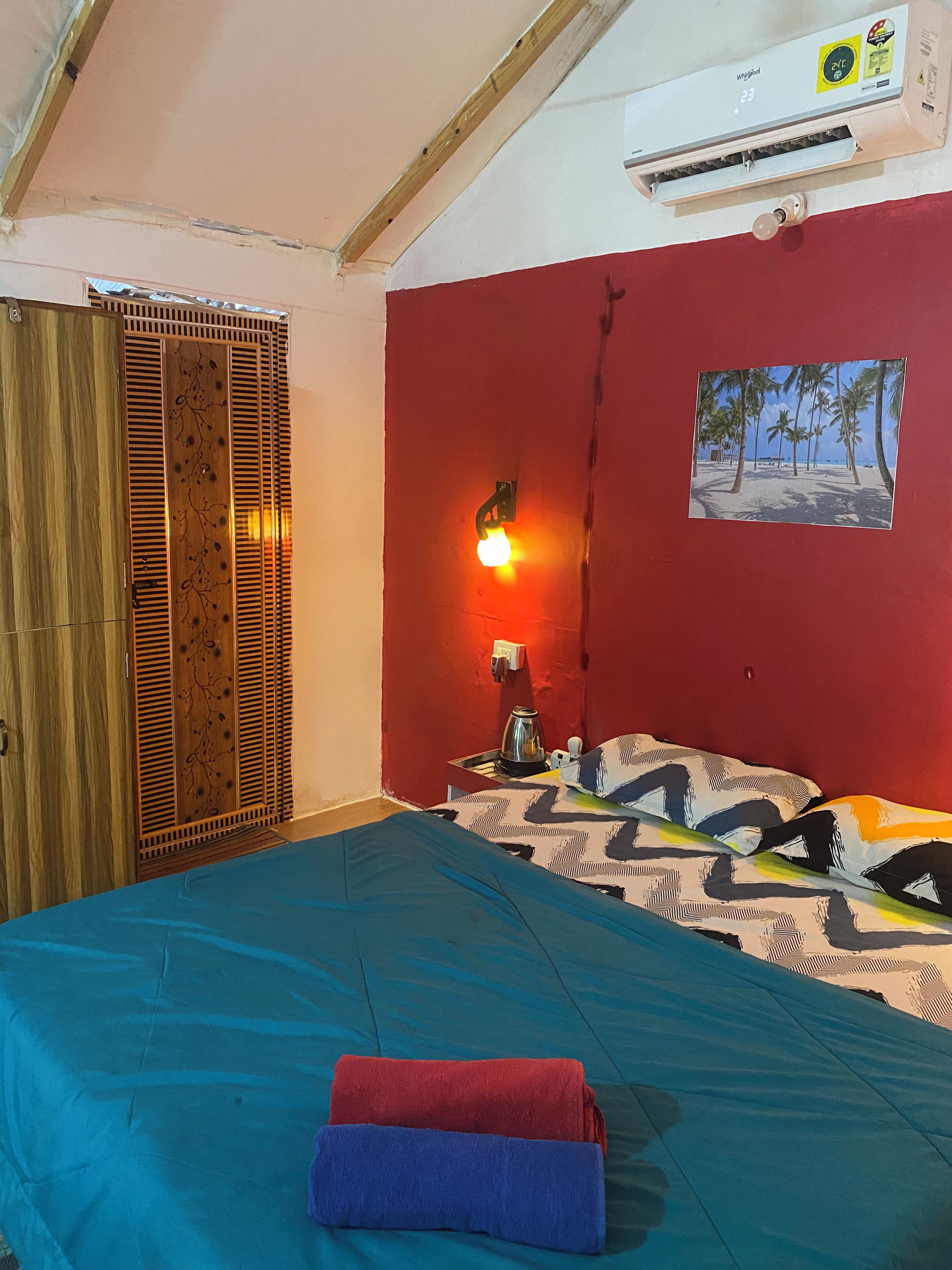 A bedroom with a bed, a red wall, a painting of a beach, and an air conditioner.