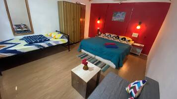 Una habitación con dos camas, un sofá, una mesita con un florero y una pared roja con un cuadro.