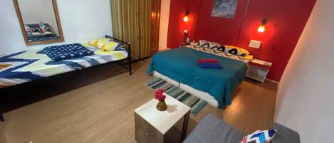 A room with two beds, a sofa, a small table with a vase, and a red wall with a picture.