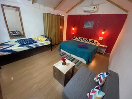Una habitación con dos camas, un sofá, una mesita con un florero y una pared roja con un cuadro.