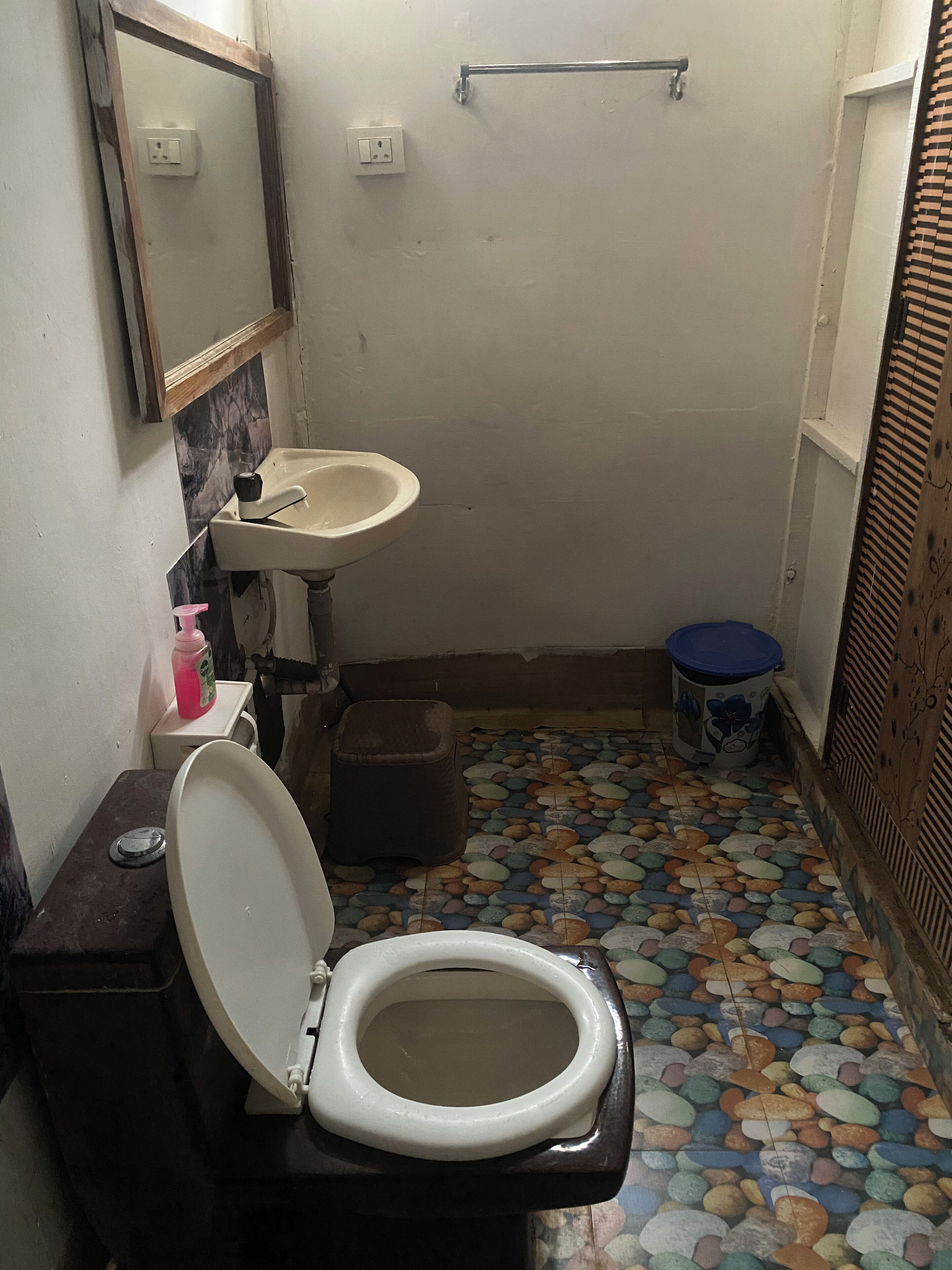 Une petite salle de bain avec des toilettes, un lavabo et un miroir. Un porte-serviettes est fixé au mur, et il y a une poubelle. Le sol est recouvert de carreaux aux motifs colorés.