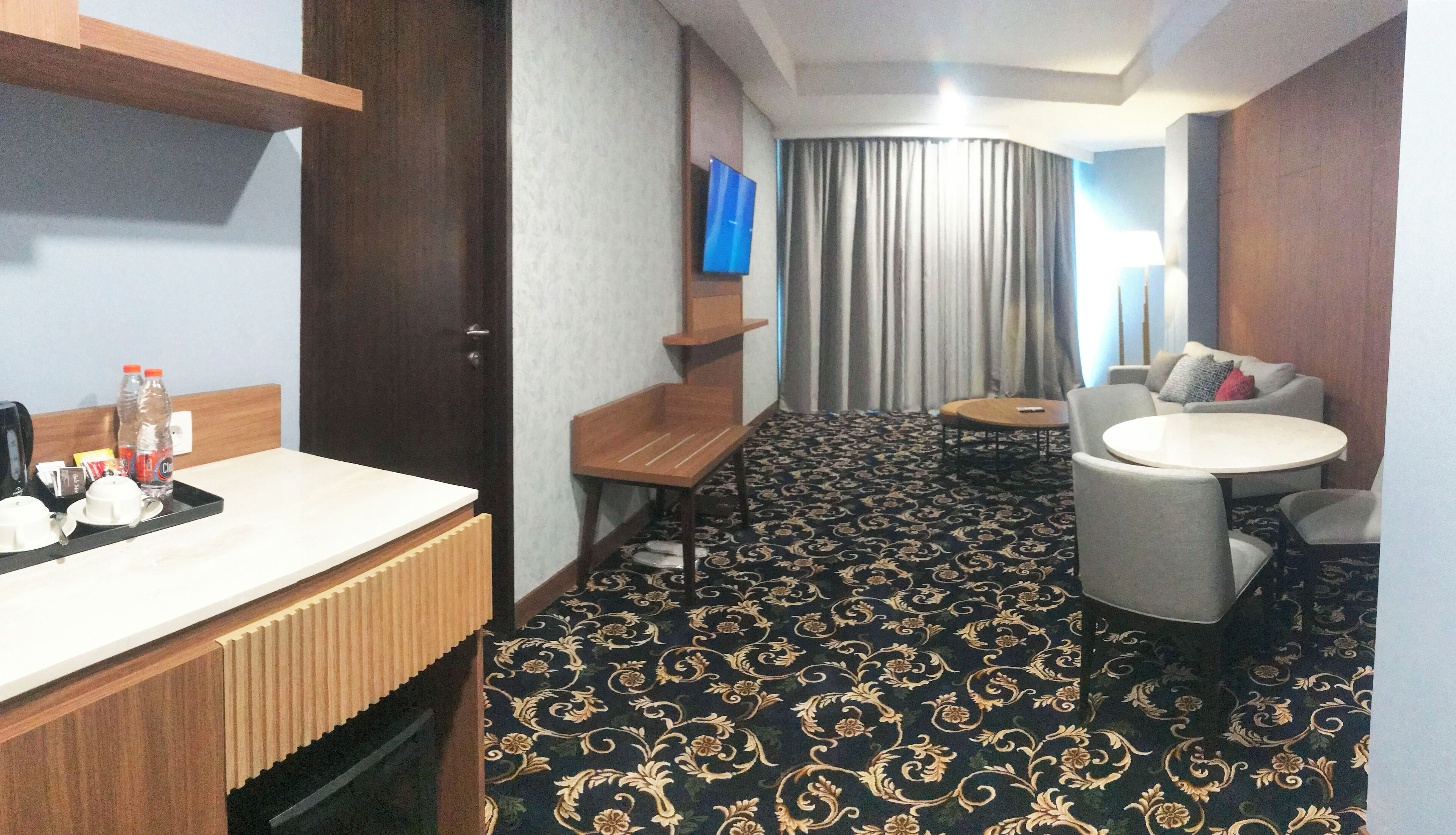 A hotel room with a wooden desk, a small round table, two chairs, a sofa, a TV, and a patterned carpet.