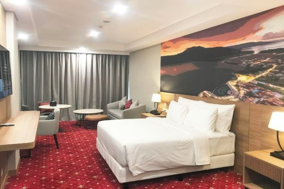 A hotel room with a large bed, a desk, a chair, a small table, and a painting of a landscape on the wall.