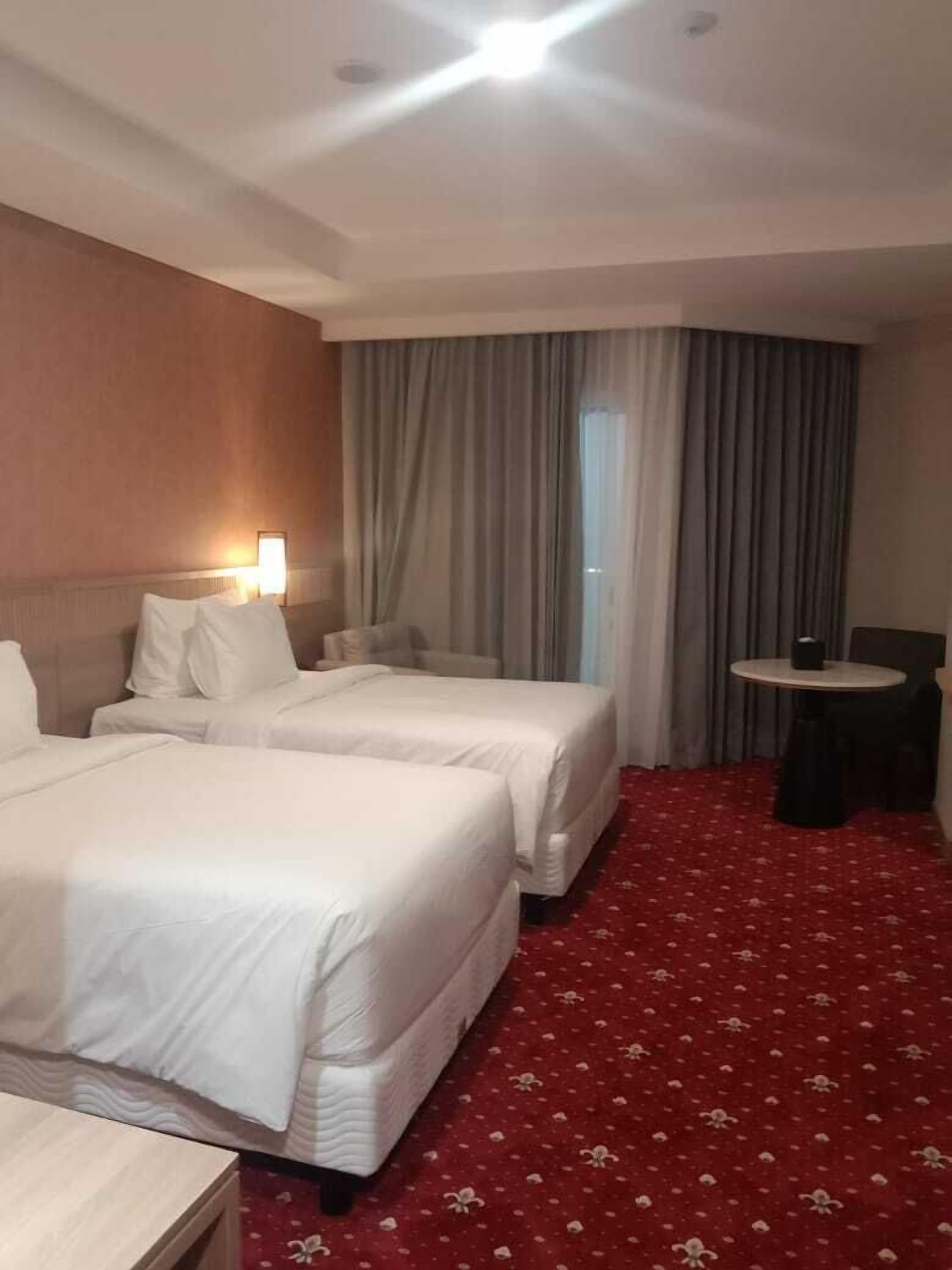 A hotel room with two beds, a small table, a lamp, and a view of the outdoors through a window with curtains.