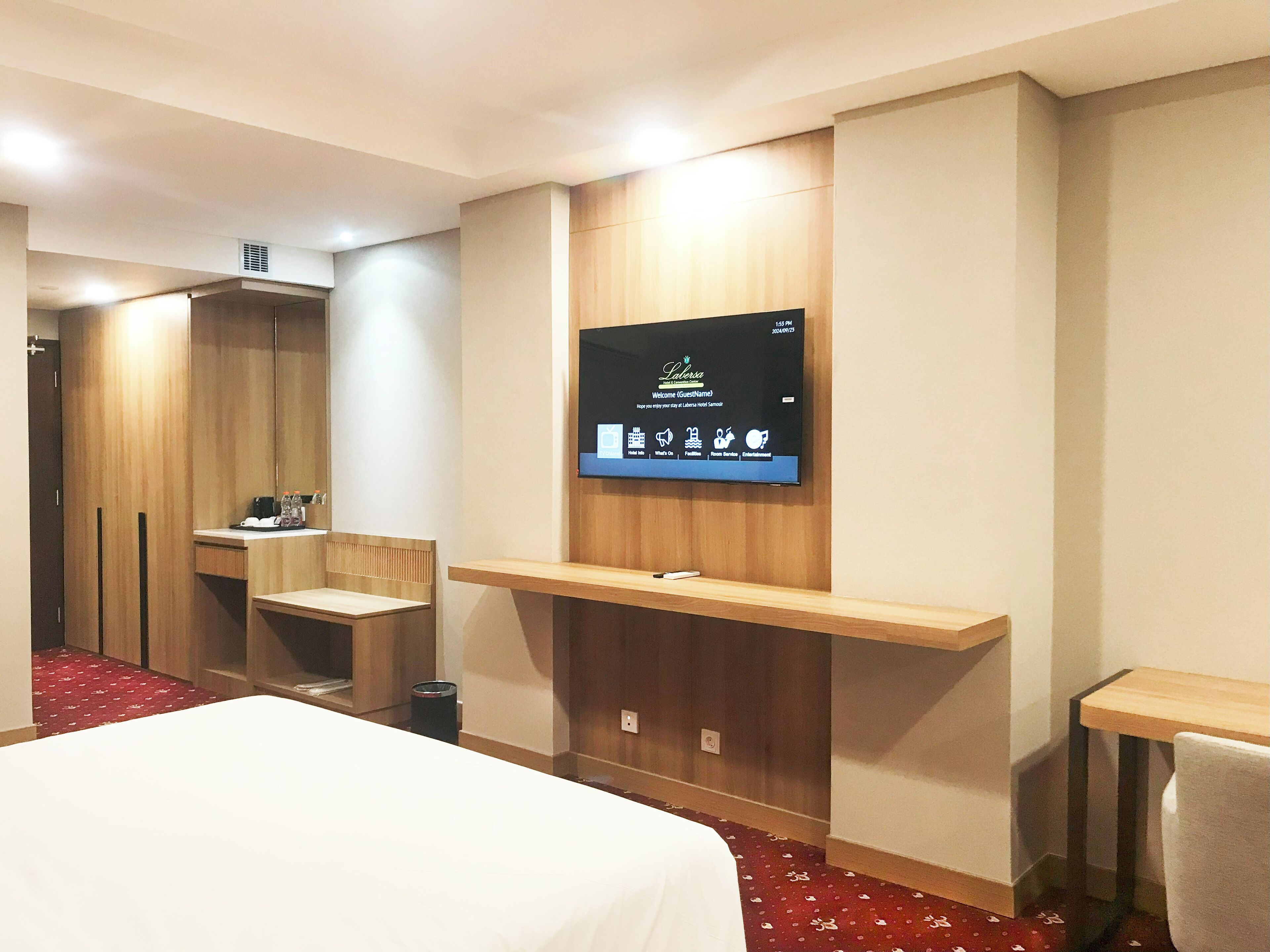 A hotel room with a bed, a TV mounted on the wall, a wooden desk, and a small seating area.