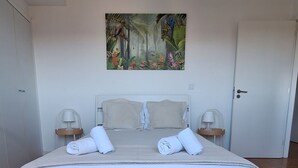 Une chambre moderne avec un grand lit, du linge de lit blanc, un luminaire suspendu et un tableau aux motifs tropicaux accroché au mur.