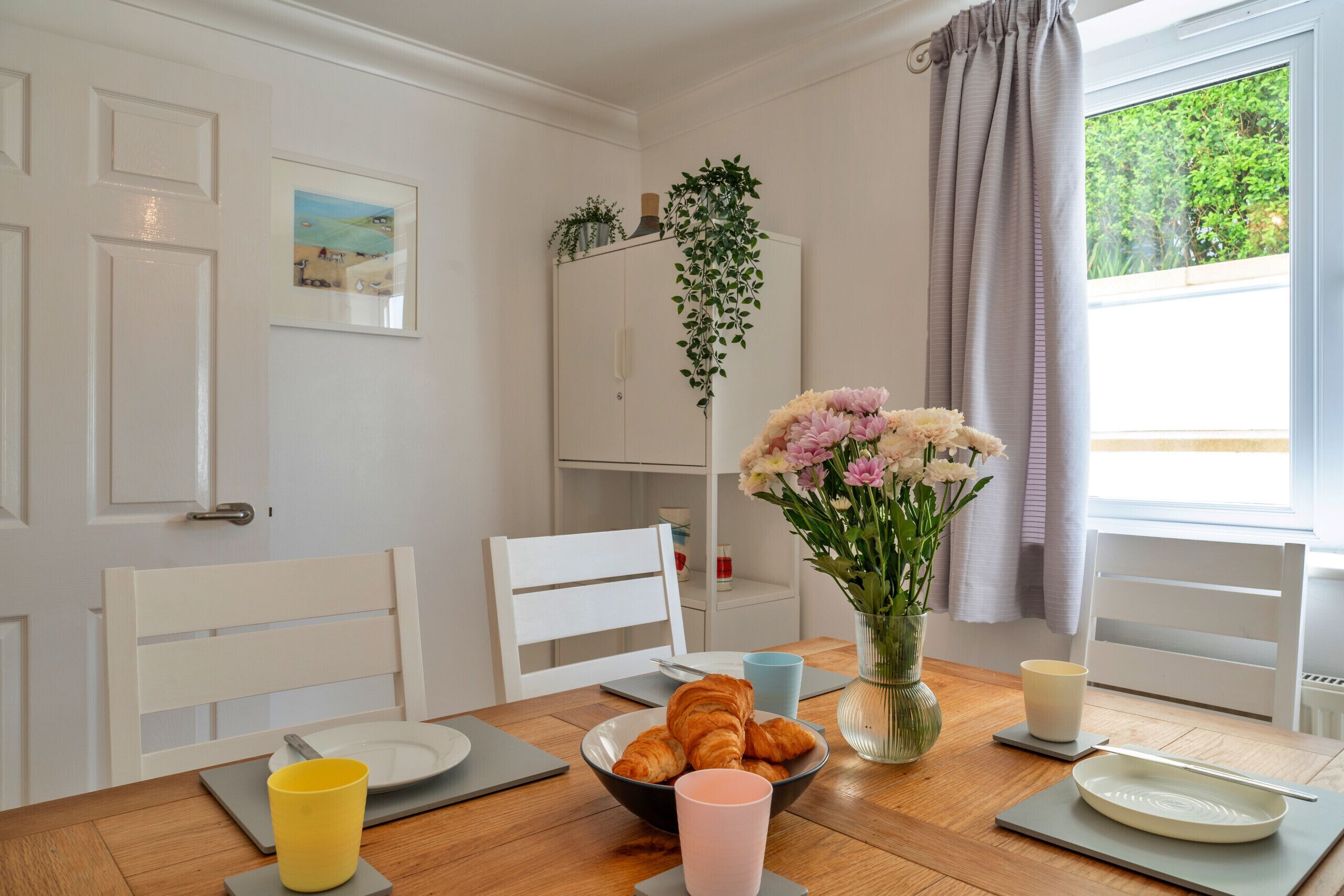 Een eethoek met een houten tafel, stoelen, een bloevas en een raam met gordijnen.