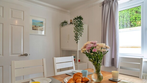 A dining area with a wooden table, chairs, a vase of flowers, and a window with curtains. - Blue Horizon (St Ives)