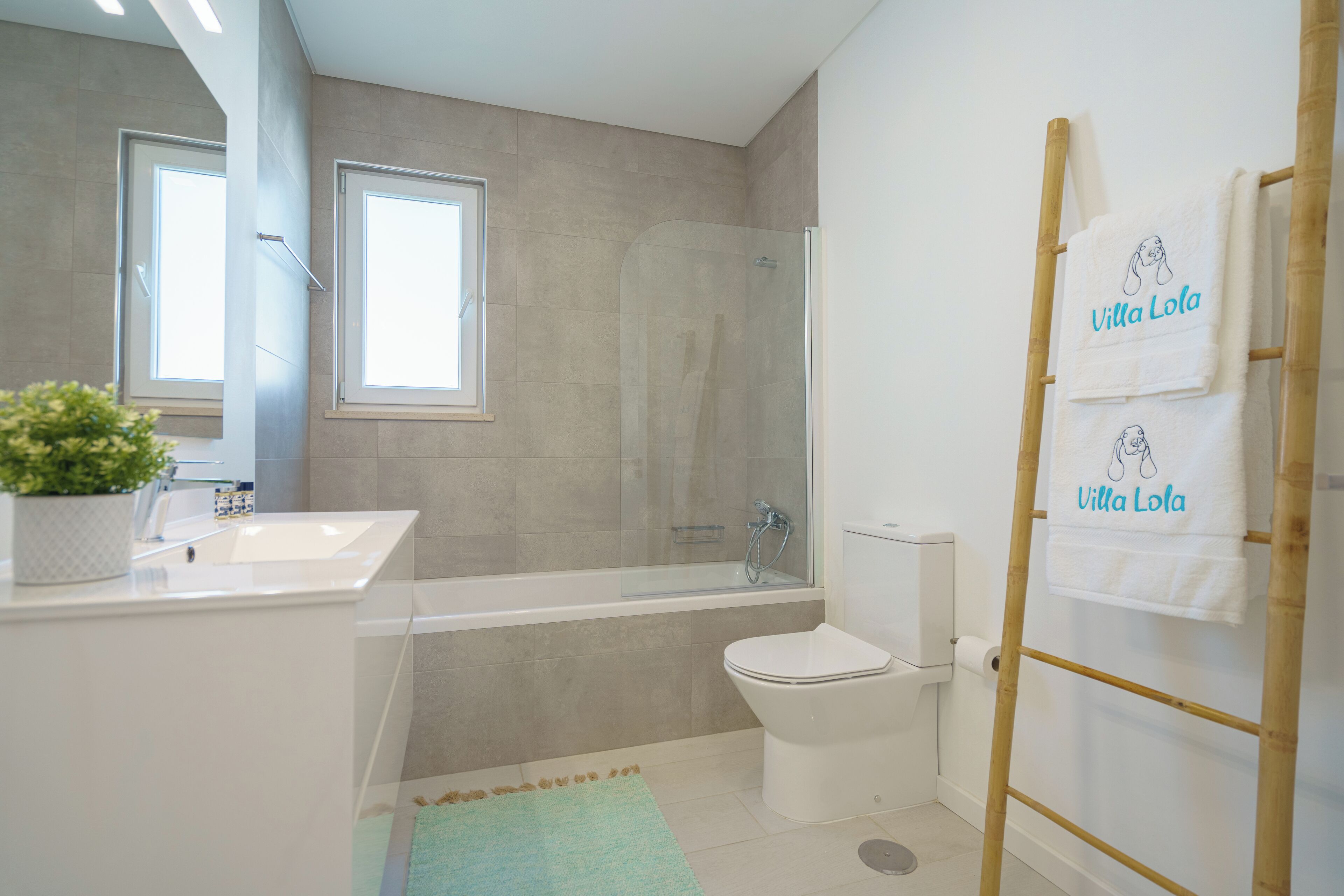 Une salle de bain moderne équipée d’une baignoire, de toilettes et d’une douche à parois de verre. Deux fenêtres laissent entrer la lumière naturelle, et une échelle en bambou sert à suspendre les serviettes.