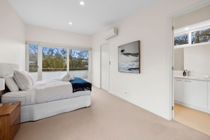 A bedroom with a bed, a bedside table, a window with a view, and a bathroom with a sink and mirror. - Yarringa Beach House (Fairhaven)