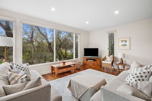 A spacious living room with a large window, a flat-screen TV, a wooden coffee table, and a comfortable sofa set. - Yarringa Beach House (Fairhaven)