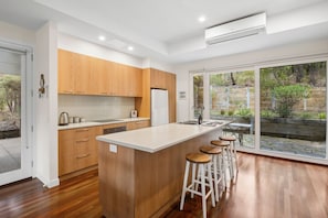 A modern kitchen with wooden cabinets, a central island with stools, and a large sliding glass door leading to an outdoor area. - Yarringa Beach House (Fairhaven)