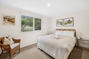 A bedroom with a large bed, a wicker chair, two framed pictures, and a window with a view of greenery.