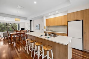 A modern kitchen with wooden cabinets, a large island with stools, and a view of trees through the window. - Yarringa Beach House (Fairhaven)