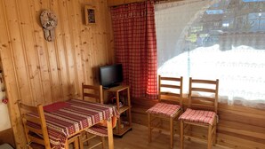 A cozy room with wooden walls, a wooden table with a checkered tablecloth, two wooden chairs, a television on a wooden stand, and a window with red checkered curtains.