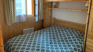 A wooden cabin room with a bed, a window with curtains, a shelf with a vase, and a wooden headboard.